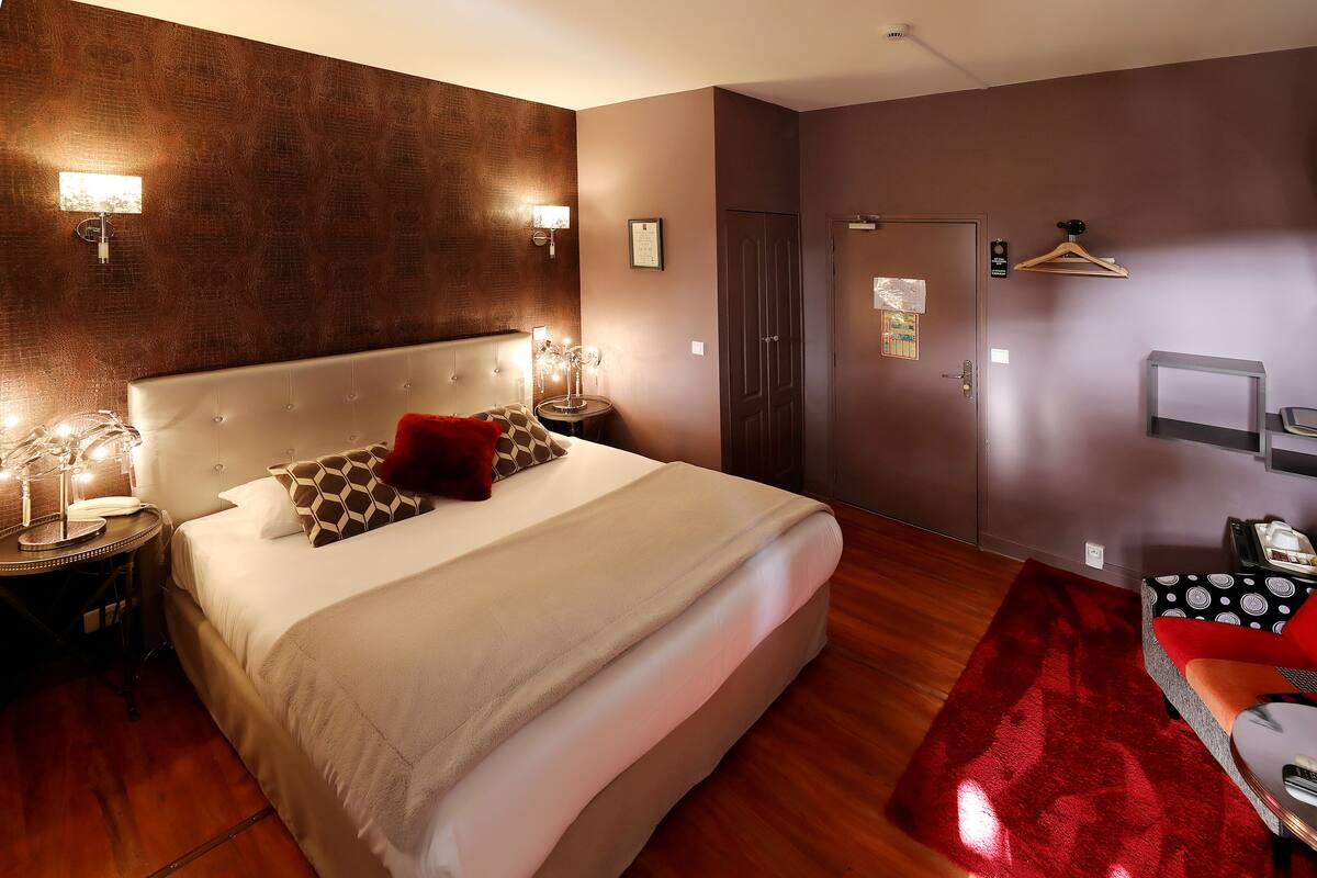 A hotel room with a large bed, bedside tables with lamps, a red rug, and a seating area with a patterned sofa.