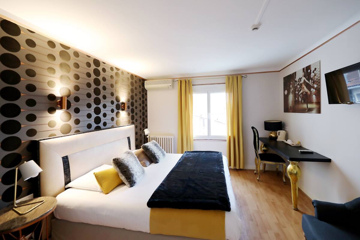 A hotel room with a bed, a desk, a chair, a lamp, and a wall with circular patterns.