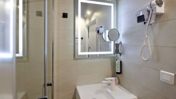 A modern bathroom with a glass shower enclosure, a white sink, a mirror with integrated lighting, and a hairdryer mounted on the wall.