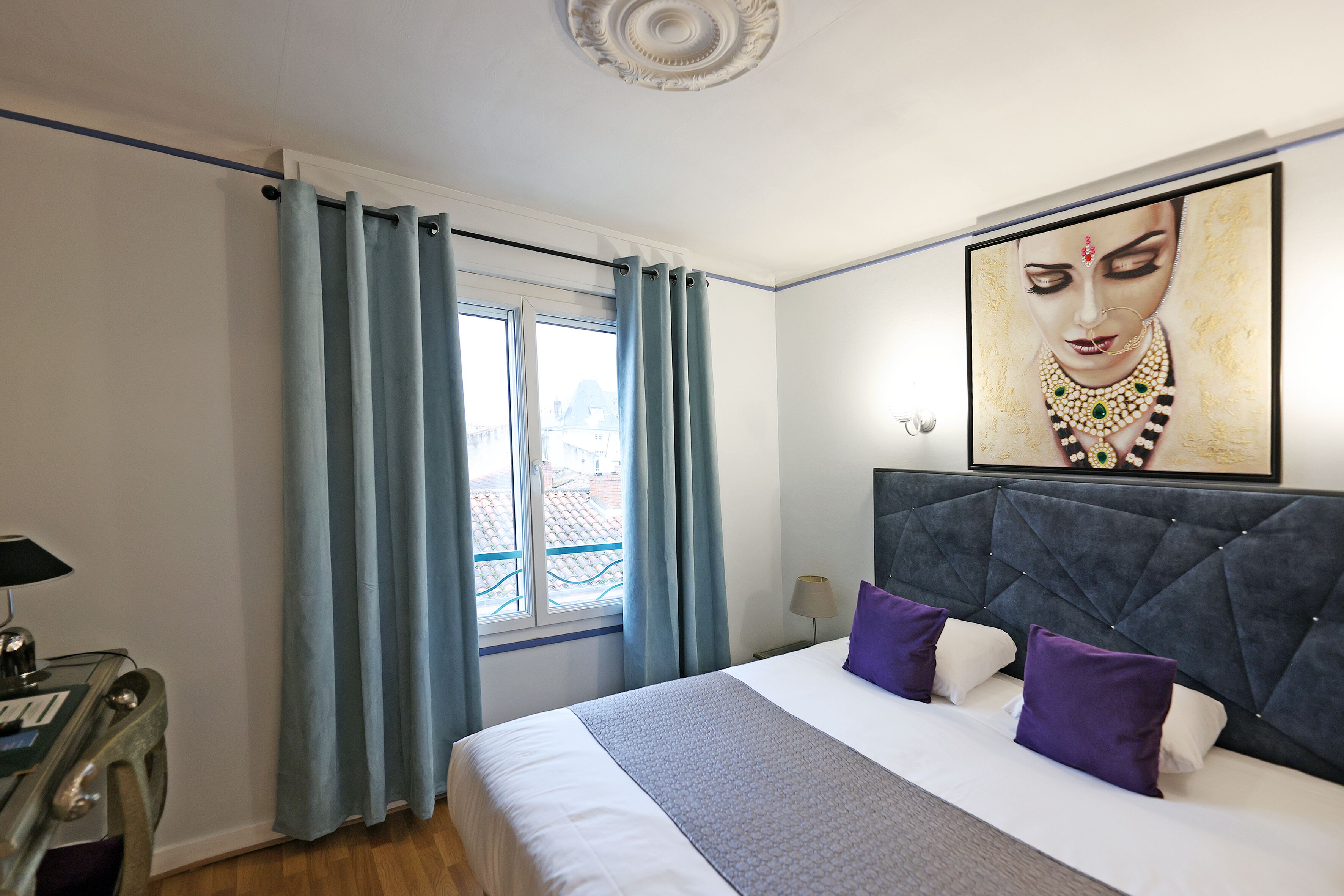 Una habitación de hotel con una cama grande, un cabecero, una ventana con cortinas, un escritorio con lámpara y un cuadro en la pared.