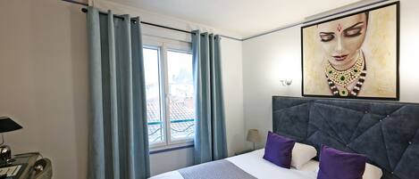 Una habitación de hotel con una cama grande, un cabecero, una ventana con cortinas, un escritorio con lámpara y un cuadro en la pared.