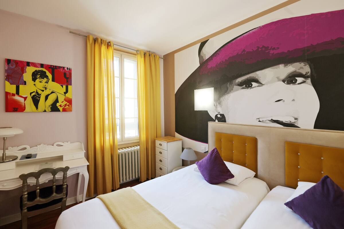 A hotel room with a bed, a desk, a chair, a radiator, a window with yellow curtains, and a large wall mural.