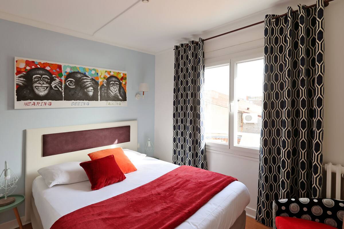 A neatly arranged bedroom with a bed, a red throw blanket, and a window with patterned curtains.