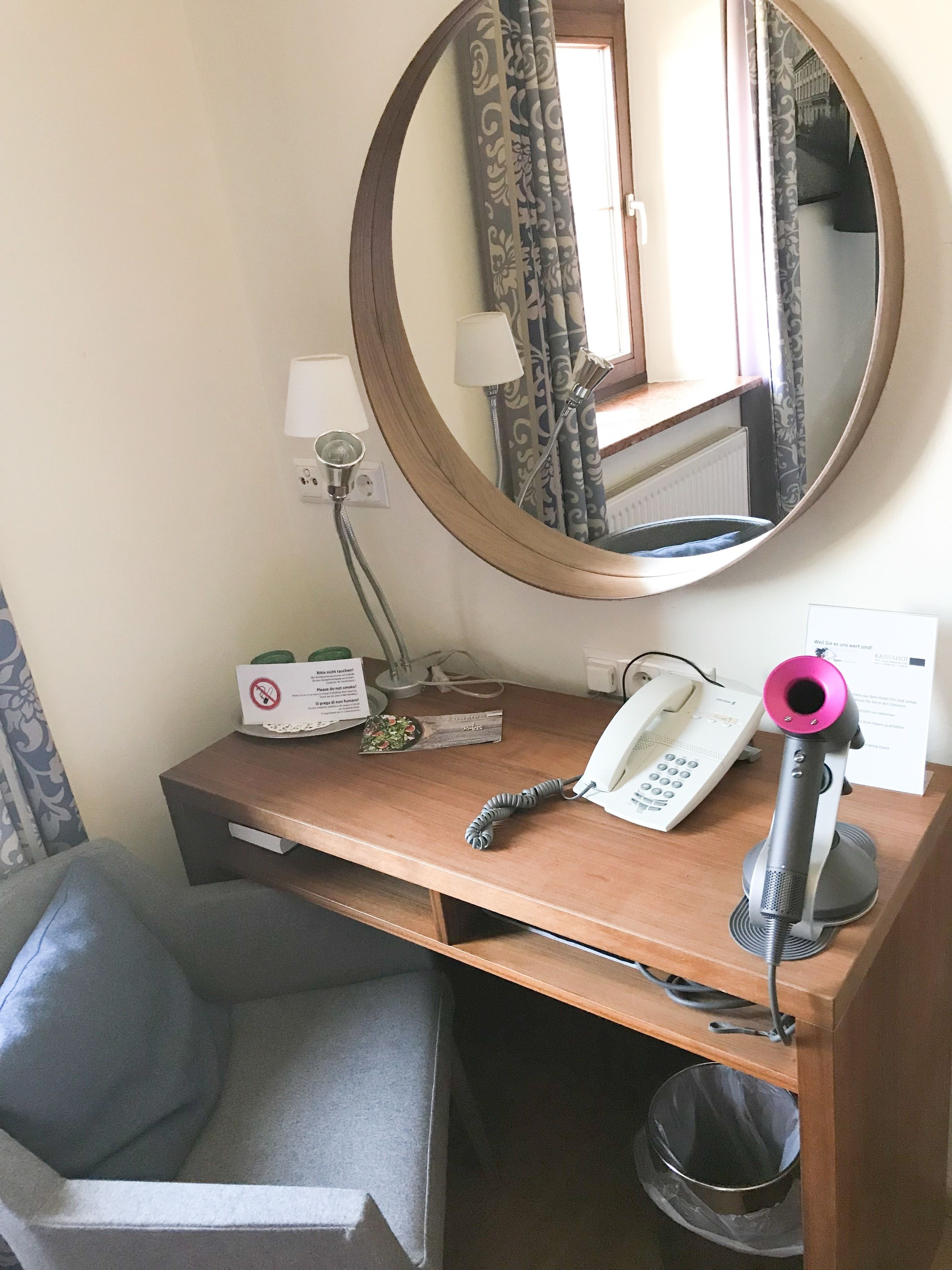 A hotel room with a wooden desk, a telephone, a hairdryer, a mirror, a lamp, and a trash bin.