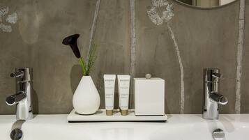 A bathroom with a white sink, two chrome faucets, a vase with a black flower, and two skincare products.