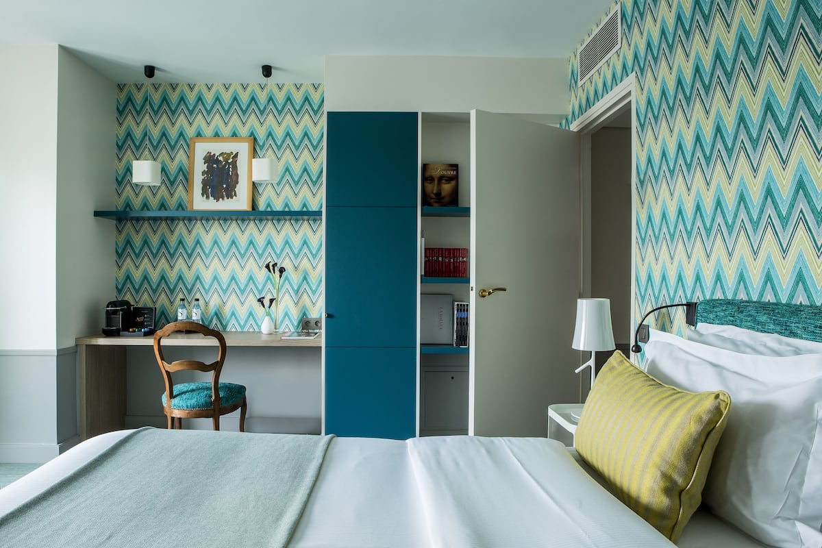 A hotel room with a bed, a desk, and a chair. There is a bookshelf with books and a picture frame.
