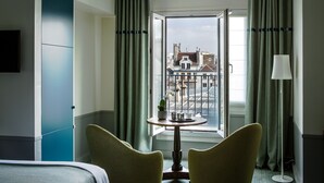 Habitación de hotel con cama, dos sillas, una mesita con una planta, una lámpara y vistas a edificios a través de un ventanal grande.