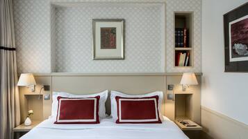 Een hotelkamer met een opgemaakt bed, twee nachtkastjes met lampen, een ingelijst schilderij aan de muur, een boekenplank en een nachtkastje met een vaas.