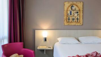 Habitación de hotel con cama, mesita de noche, lámpara, silla y una obra de arte en la pared.