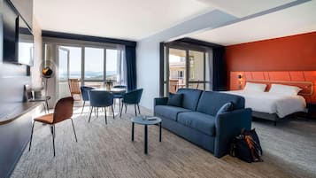 Habitación de hotel con cama, sofá, mesa de comedor con sillas, escritorio con silla, ventilador de pie y vistas al exterior.