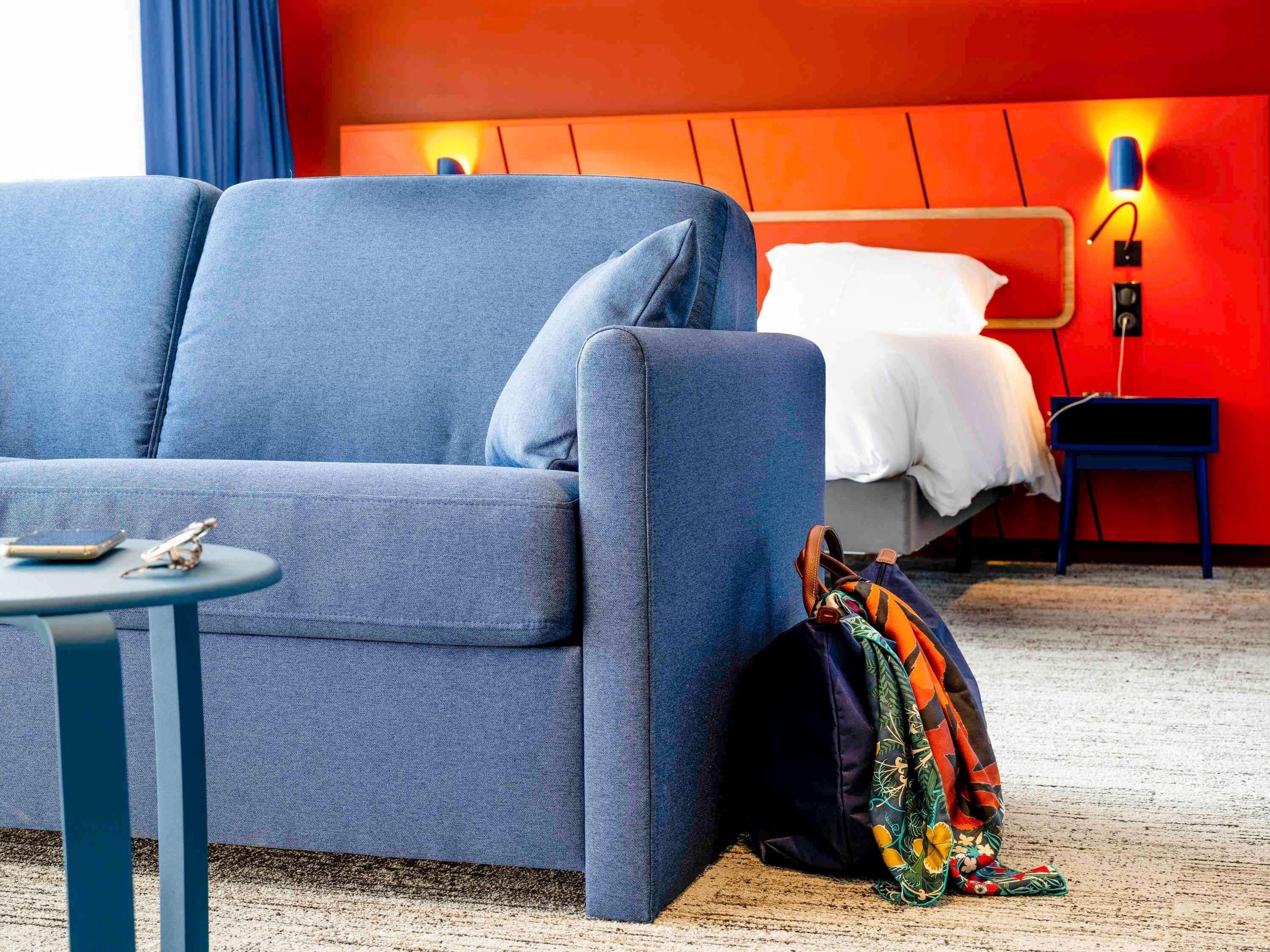 Una habitación de hotel moderna con un sofá azul, una mesita con un teléfono y un bolígrafo, una cama con ropa de cama blanca, un bolso colorido y una pared roja con una lámpara.