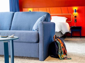 Quarto de hotel moderno com sofá azul, uma mesinha com telefone e caneta, cama com roupa de cama branca, uma mala colorida e uma parede vermelha com luminária.