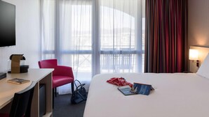 Una habitación de hotel con cama, escritorio, silla, televisor y ventana con cortinas.