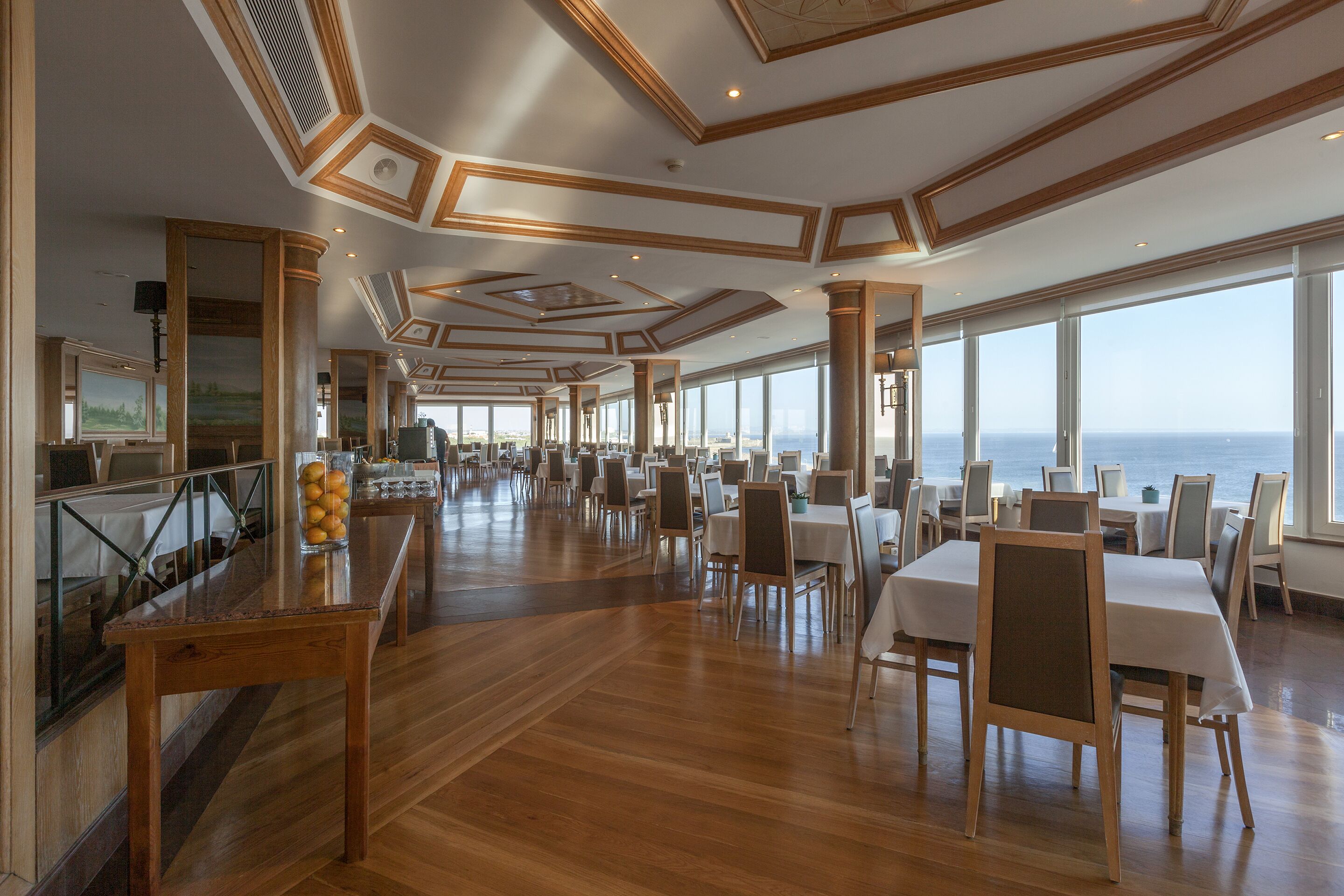 Un vaste espace dédié à la salle à manger, doté d’un plancher en bois, de grandes fenêtres offrant une vue sur l’océan et d’un bar avec un présentoir de fruits.