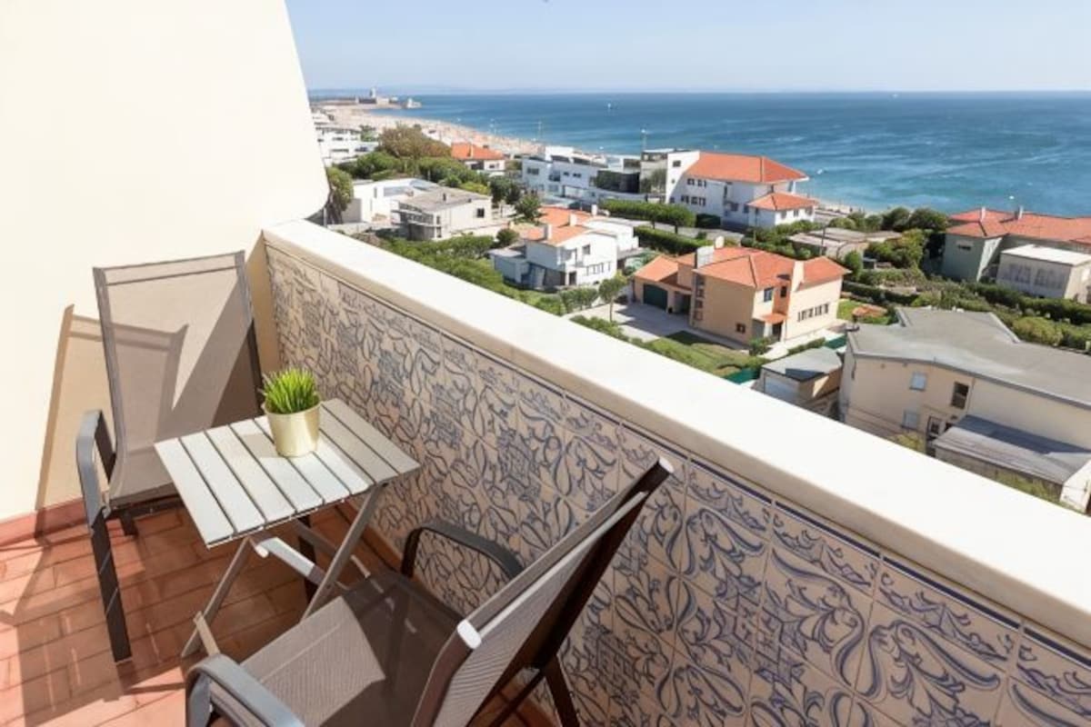 Un balcón con mesa y sillas, con vista a una zona residencial costera con casas y el mar.