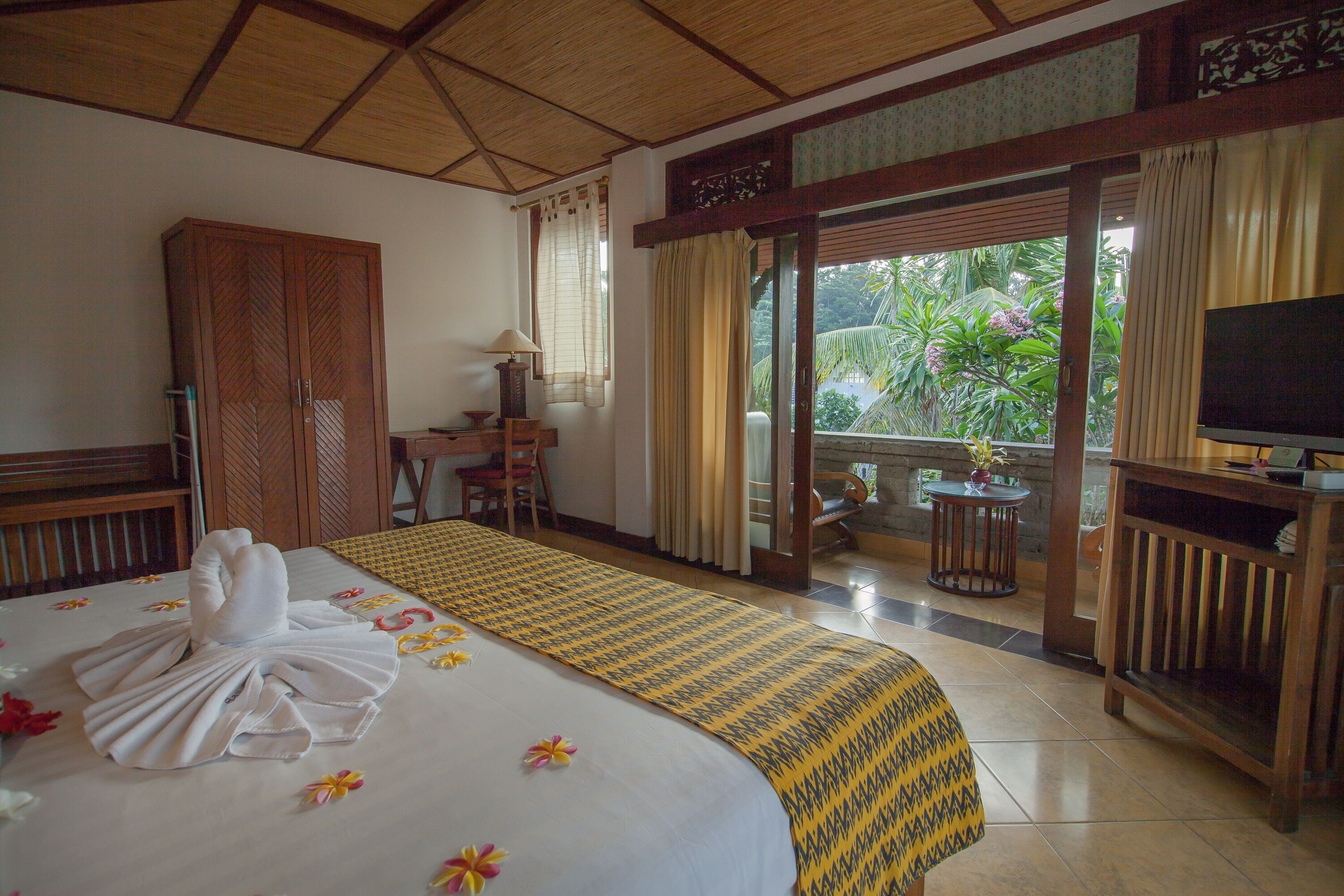 A hotel room with a bed, a desk, a television, and a balcony with a view of greenery.
