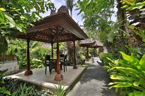 A covered outdoor seating area with tables and chairs, surrounded by lush greenery and a pathway. - Bali Spirit Hotel & Spa (Ubud)