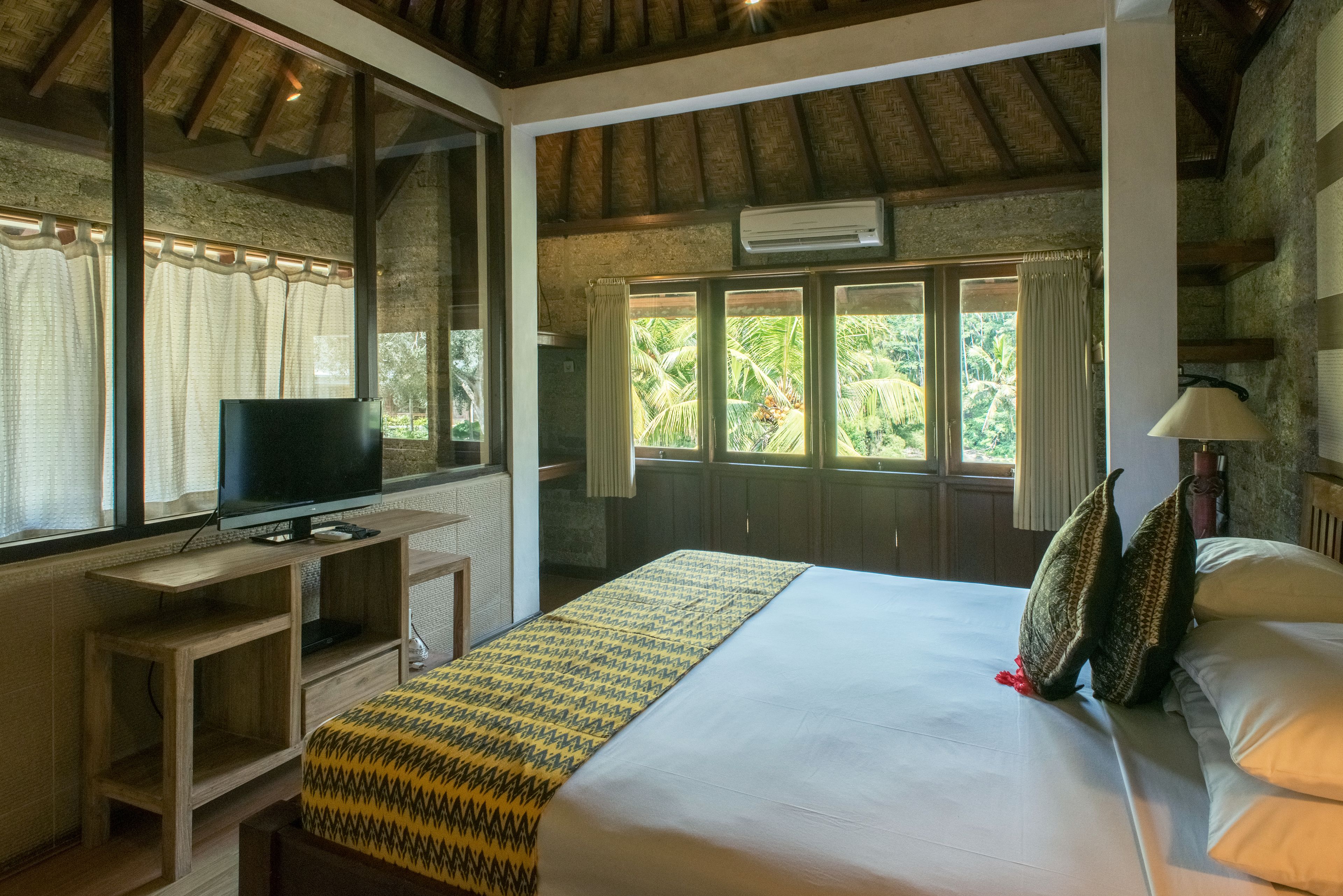 A bedroom with a large bed, a TV on a wooden stand, a window with curtains, and a view of greenery outside.