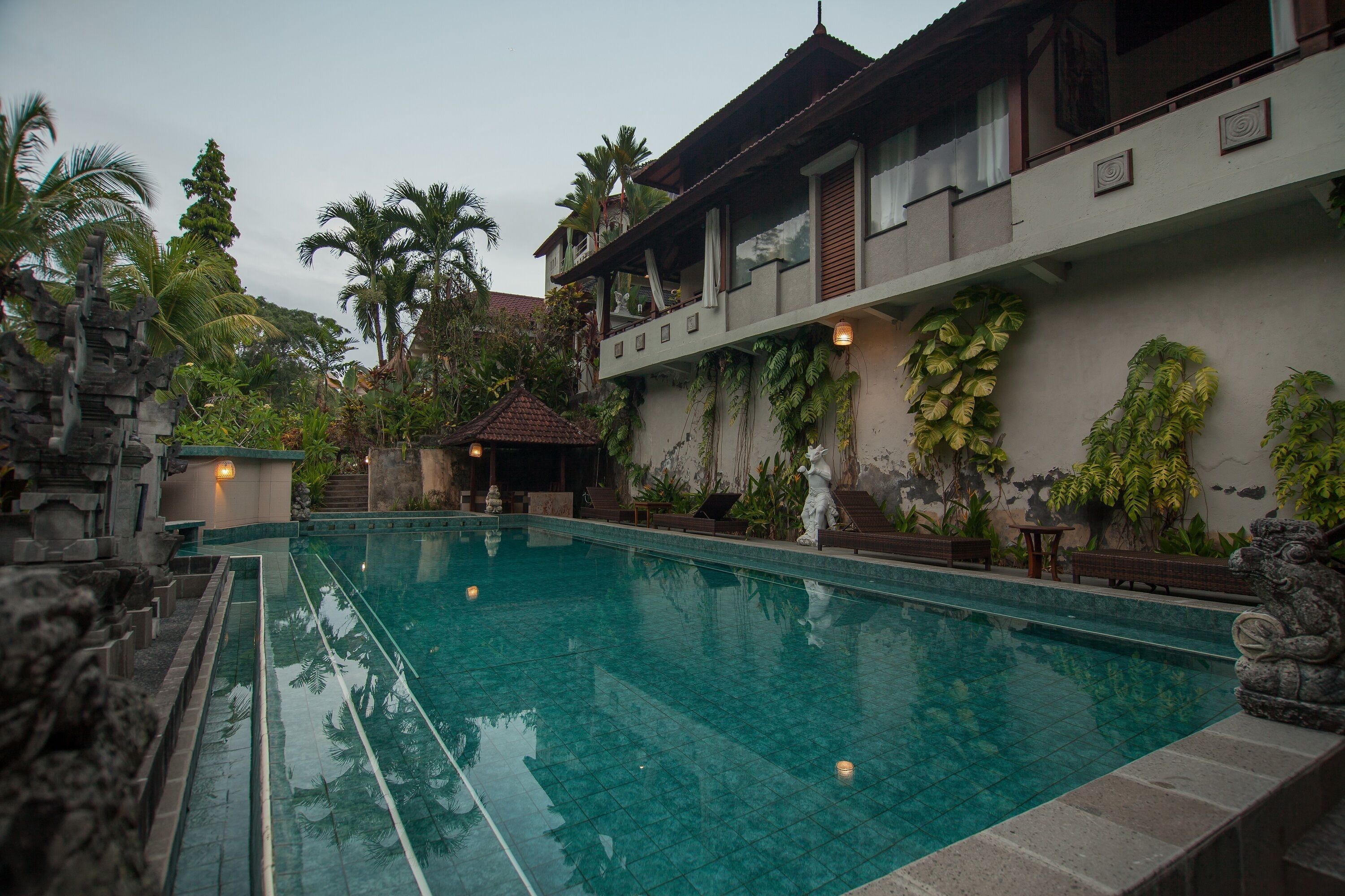 Une piscine bordée de pierres, entourée de plantes tropicales et un bâtiment traditionnel doté d’un balcon.