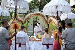 A ceremonial event with attendees seated at a table, a person performing a ritual, and a backdrop with decorative elements. - Bali Spirit Hotel & Spa (Ubud)