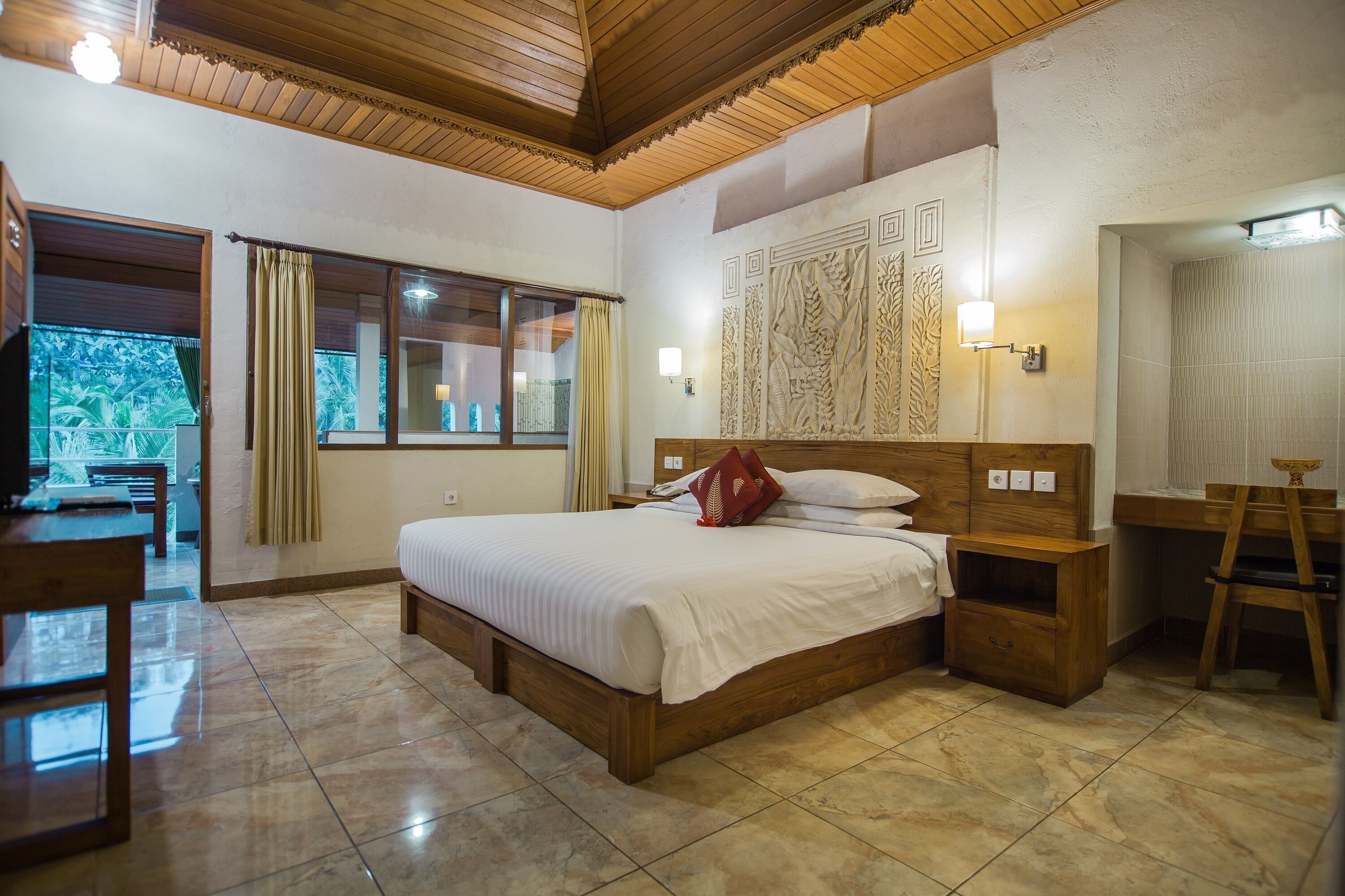 A hotel room with a wooden bed, a desk, a chair, a bathroom with a mirror, and a view of the outdoors.