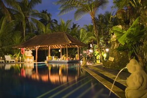 A pool area with a covered seating area, palm trees, and a stone frog statue. - Bali Spirit Hotel & Spa (Ubud)