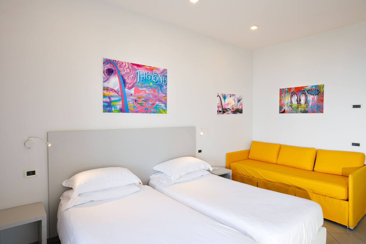 Ein Hotelzimmer mit zwei Betten, einem leuchtend gelben Sofa und farbenfroher Kunst an den Wänden.