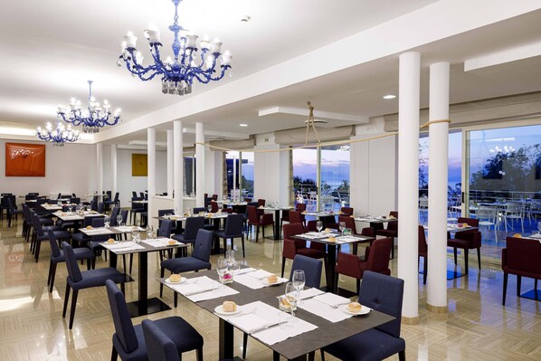 A spacious dining area with multiple tables and chairs, large windows offering a view, and elegant blue chandeliers.