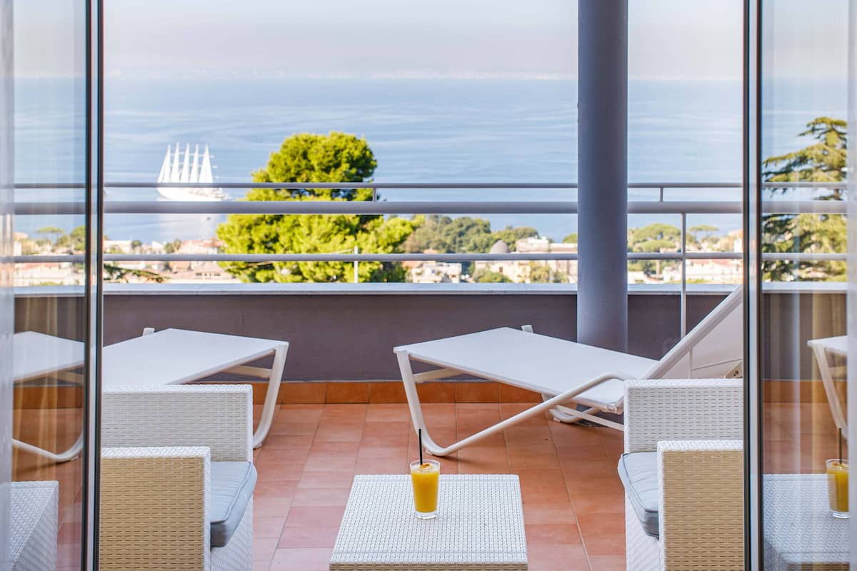 Ein Balkon mit weißen Korbmöbeln, einem Glastisch mit Orangensaft und Blick auf das Meer und ein Segelboot.
