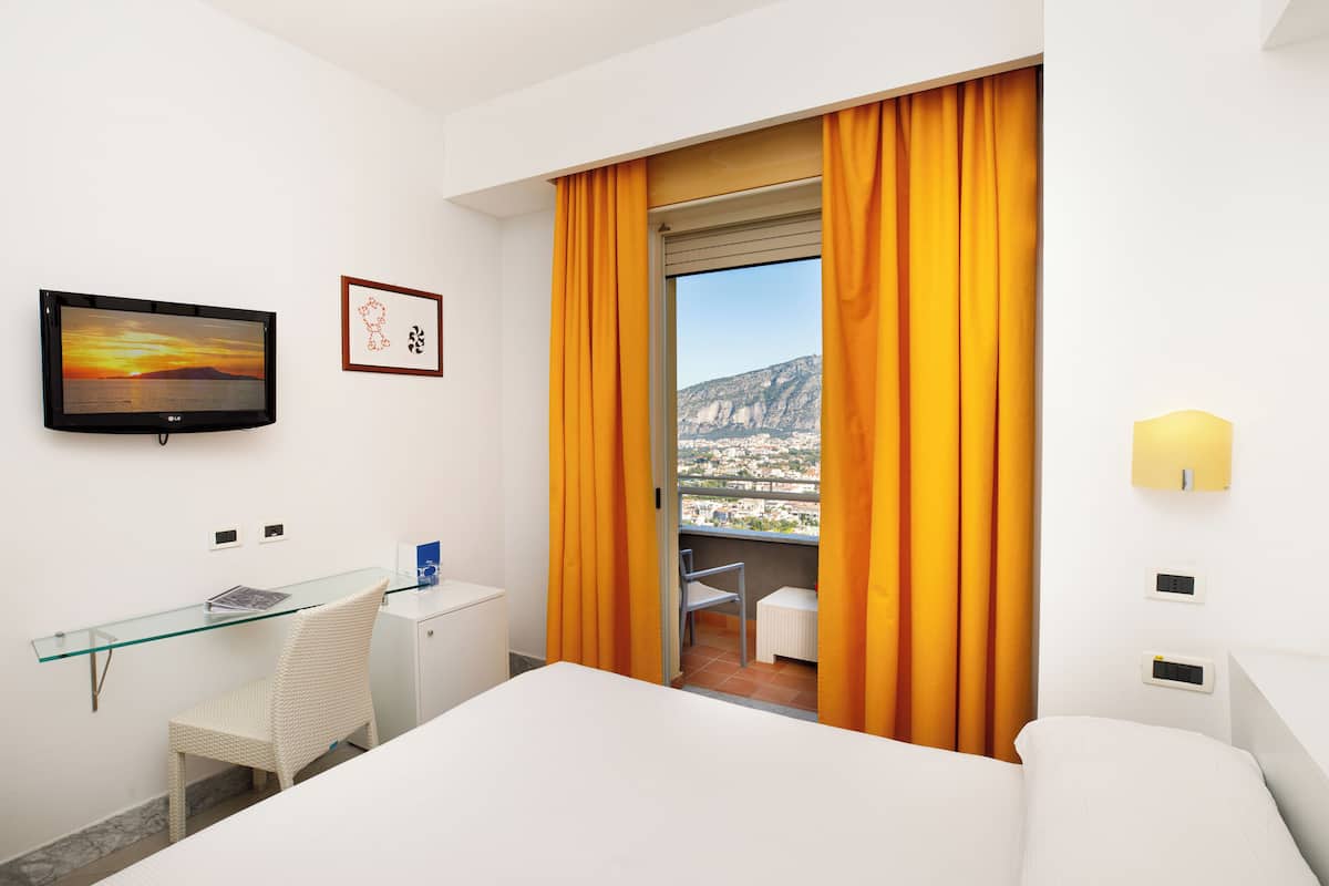 Ein Hotelzimmer mit Bett, Schreibtisch, Stuhl, Fernseher und einem Balkon mit orangenen Vorhängen.