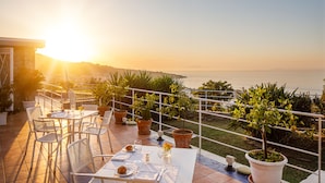 Una terraza con mesa lista para comer, plantas en macetas y vista al mar al atardecer.