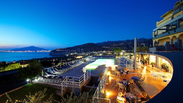Una terraza en la azotea con piscina, mobiliario exterior y vistas a la costa al atardecer.