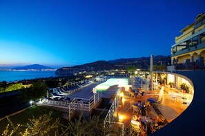 Una terraza en la azotea con piscina, mobiliario exterior y vistas a la costa al atardecer.