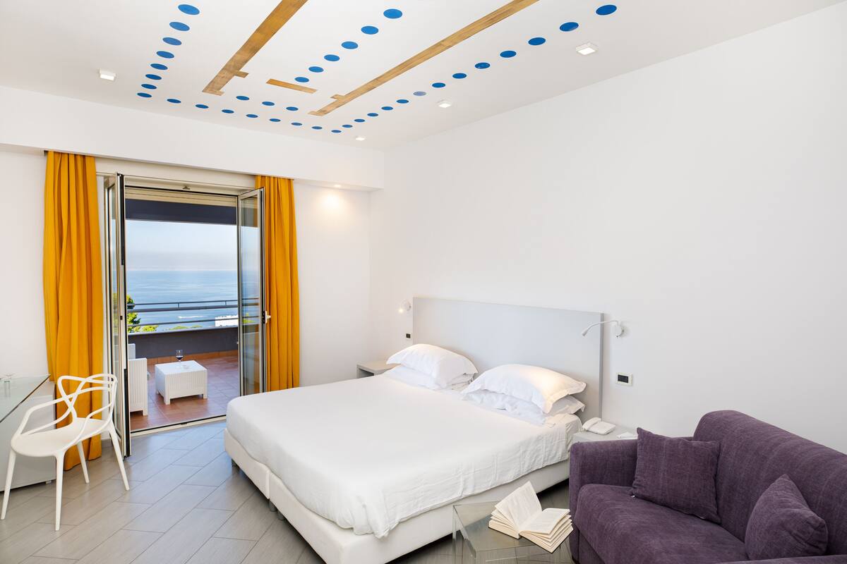 Ein modernes Hotelzimmer mit einem großen Bett, einem violetten Sofa, einem Glastisch und einem Balkon mit gelben Vorhängen.