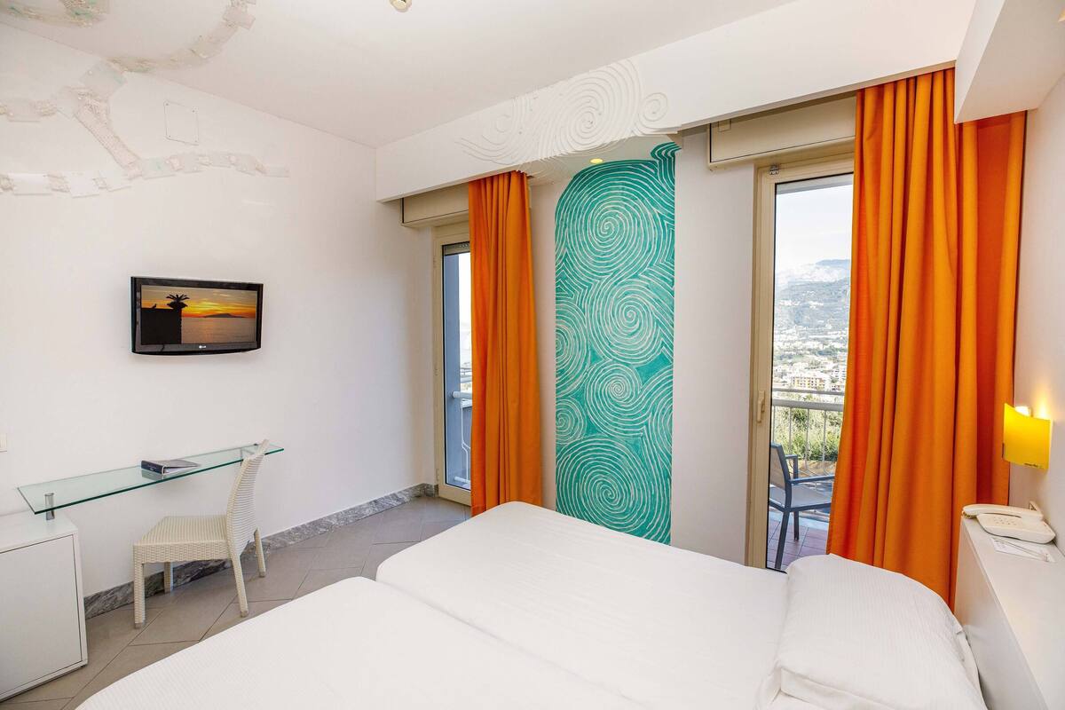 Ein Hotelzimmer mit Bett, Schreibtisch mit Stuhl, einer Glastrennwand und Blick auf die Stadt durch große Fenster.