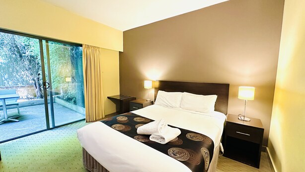 A hotel room with a large bed, two bedside tables with lamps, a sliding glass door leading to an outdoor area, and a bench outside.