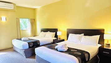 A hotel room with two beds, each with white linens and a patterned bedspread. There is a window with curtains, a wall-mounted air conditioner, and a bedside table with a lamp.