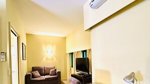 A compact living space with a sofa, a desk, a television, and an air conditioning unit.