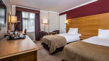 A hotel room with two beds, a desk with a lamp, a chair, and a window with curtains.