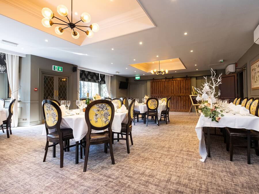 A dining area with round tables, chairs, and a chandelier. There are windows with curtains, a door with an exit sign, and a wall-mounted air conditioner.