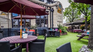 An outdoor seating area with round tables and wicker chairs, a building with a sign reading "HOTEL," and a tree in the background. - The Bridge Hotel Greenford (Greenford)