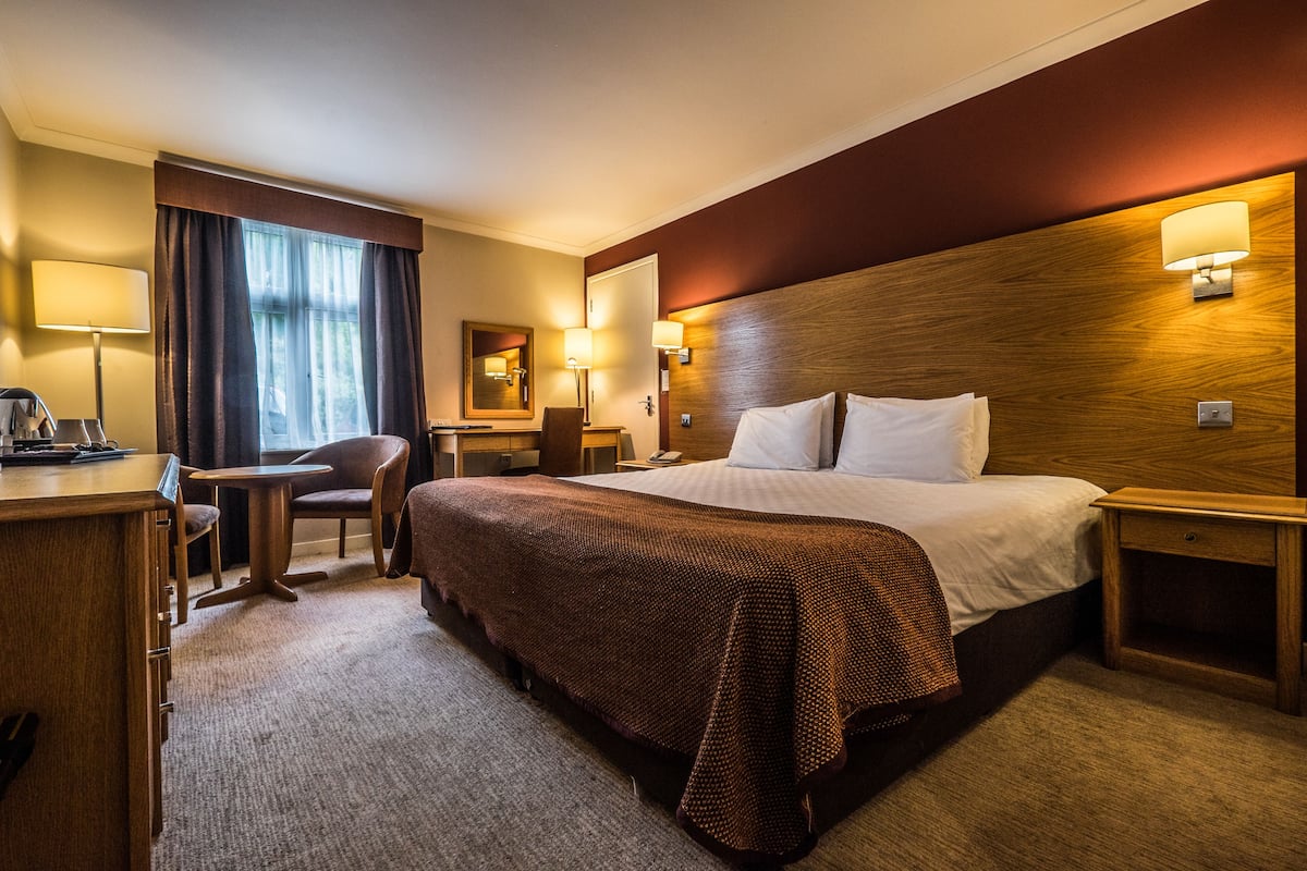 A hotel room with a large bed, a desk, a chair, a nightstand, and a lamp.