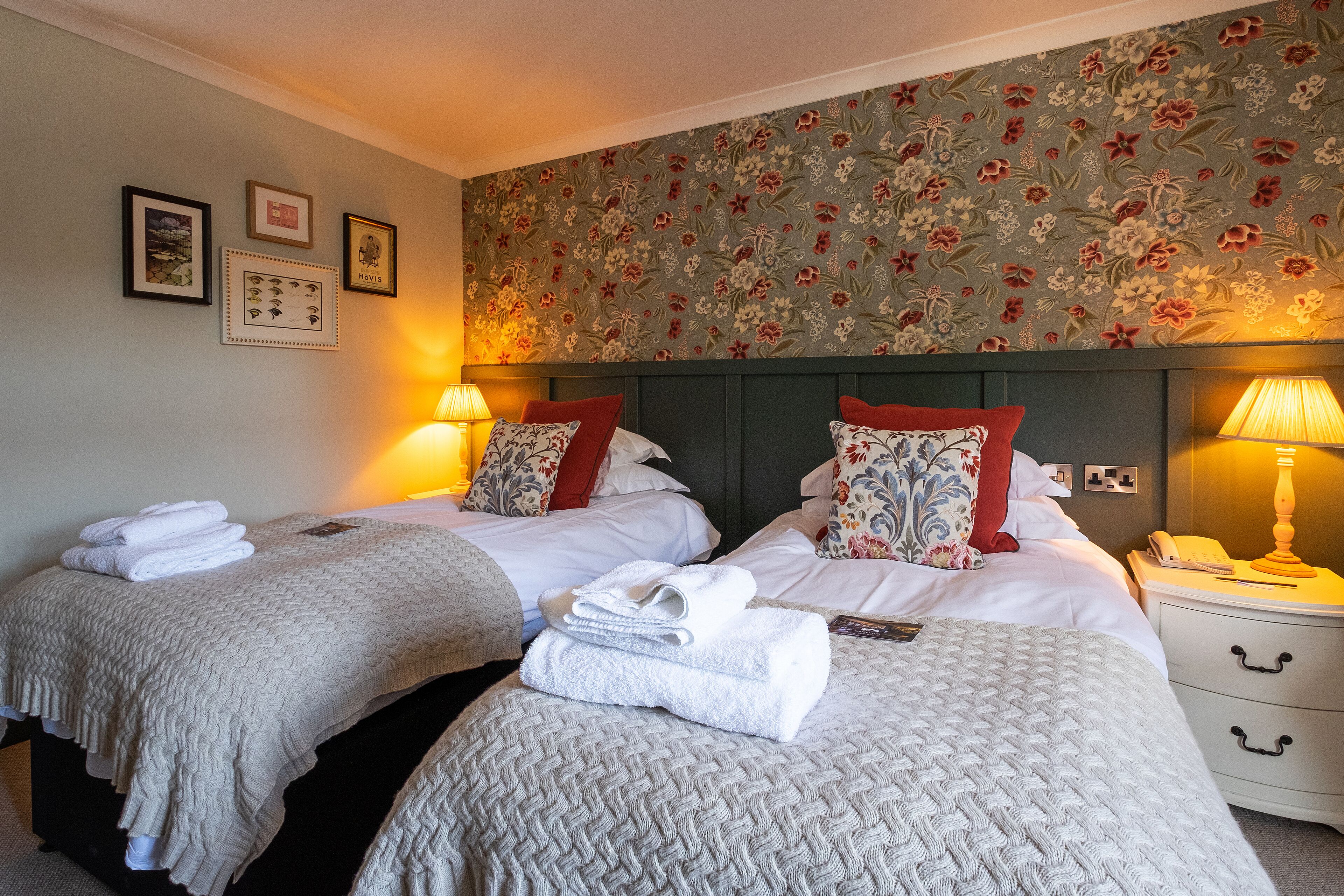 A hotel room with two beds, each with a white bedspread, red and floral pillows, and a folded towel on the bed. There are two bedside tables with lamps and a wall-mounted telephone.