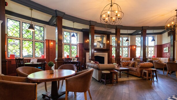 A spacious room with a fireplace, wooden flooring, and large windows with stained glass. - The Bridge Hotel Greenford (Greenford)