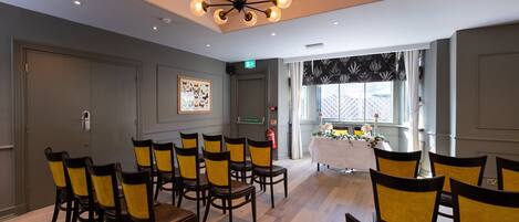 A conference room with a modern chandelier, wooden chairs, a white table with a floral arrangement, and a window with patterned curtains.