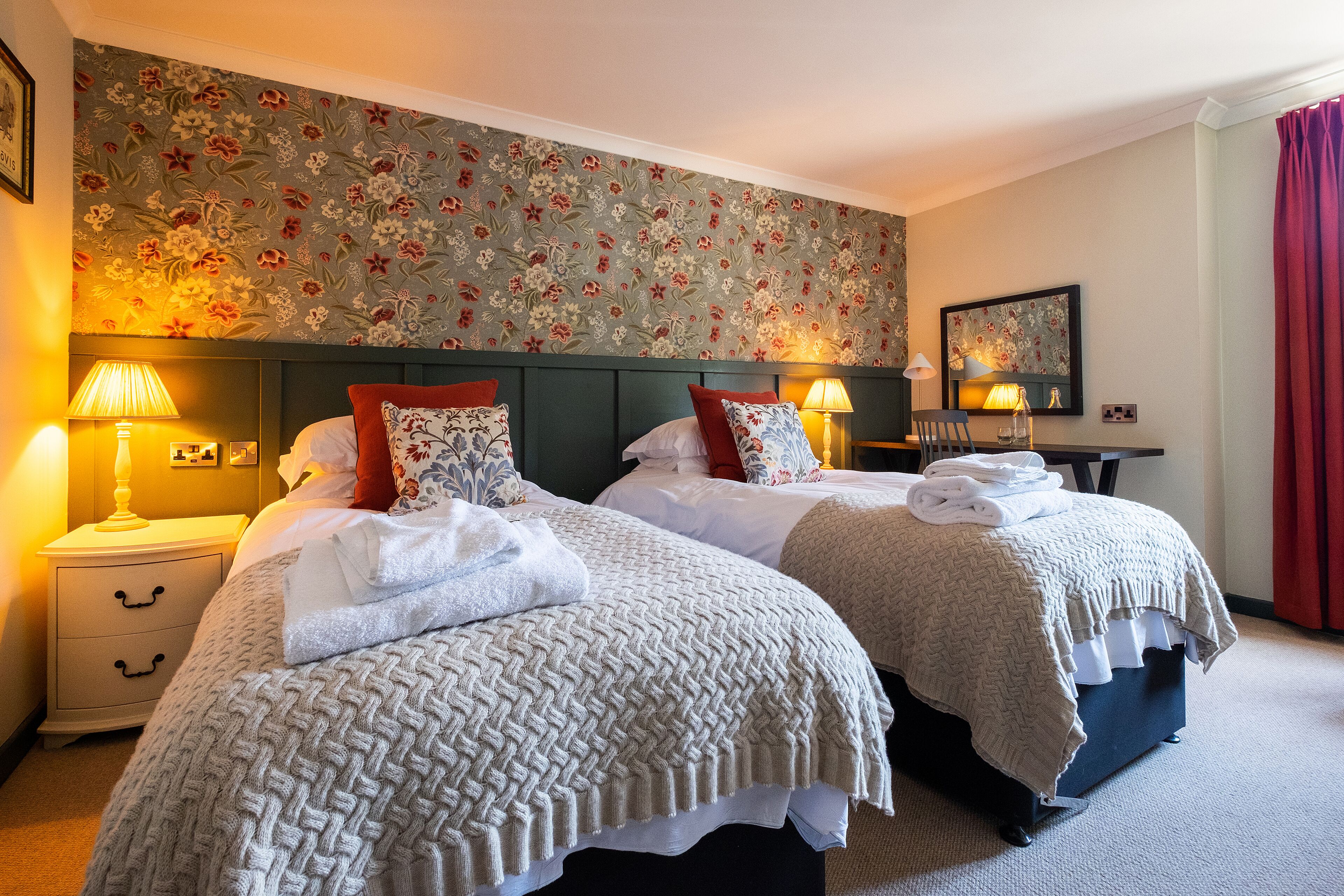 A hotel room with two beds, a nightstand with a lamp, a desk with a lamp, and a floral wallpaper.
