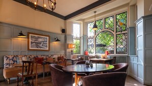 A dining area with a round table, chairs, and a bench. There are windows with decorative glass panes and a view of greenery outside. - The Bridge Hotel Greenford (Greenford)