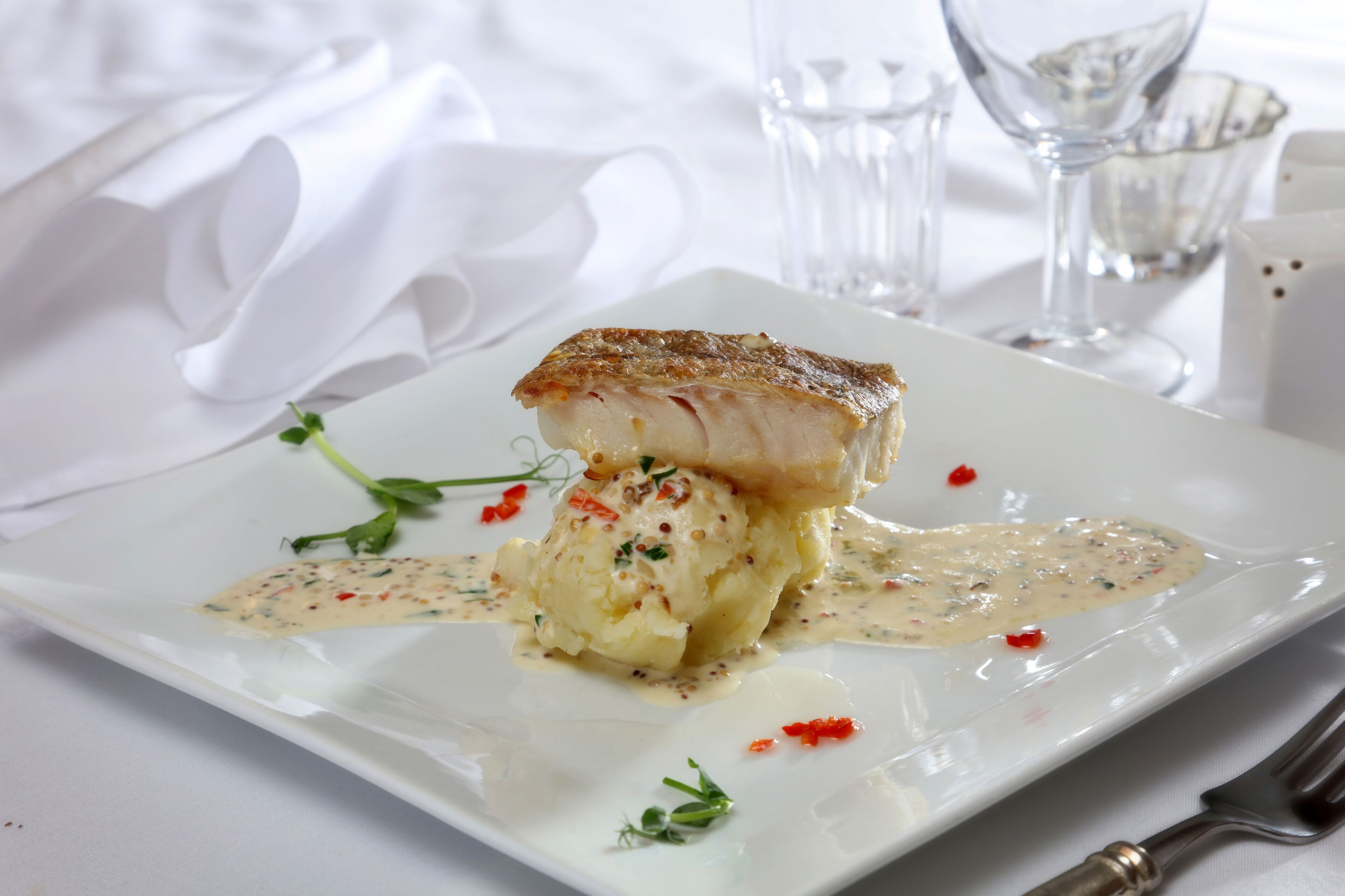 A white plate with a piece of fish, mashed potatoes, and a creamy sauce, garnished with herbs and red pepper flakes.