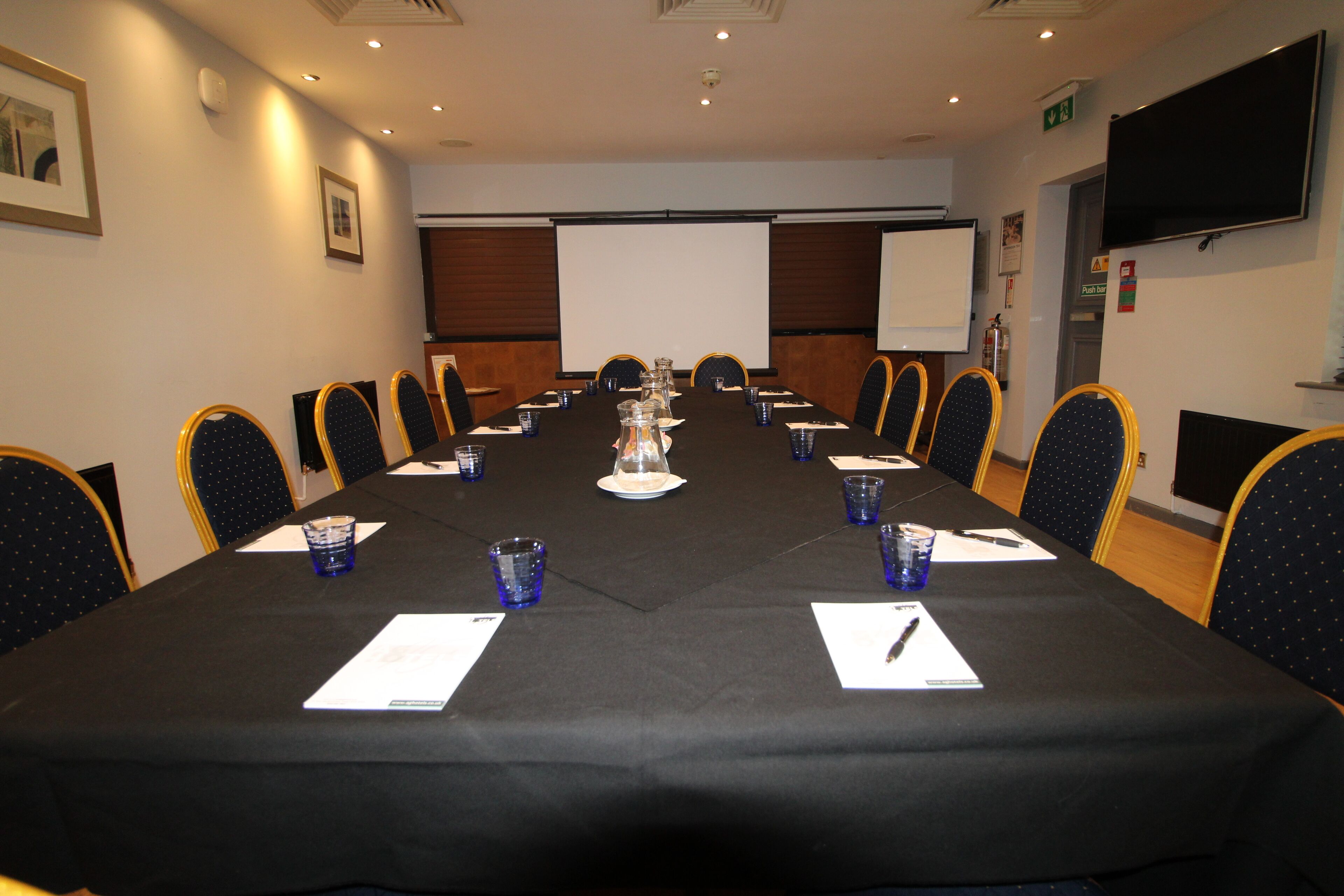A conference room with a long table, chairs, a projector screen, and a television.