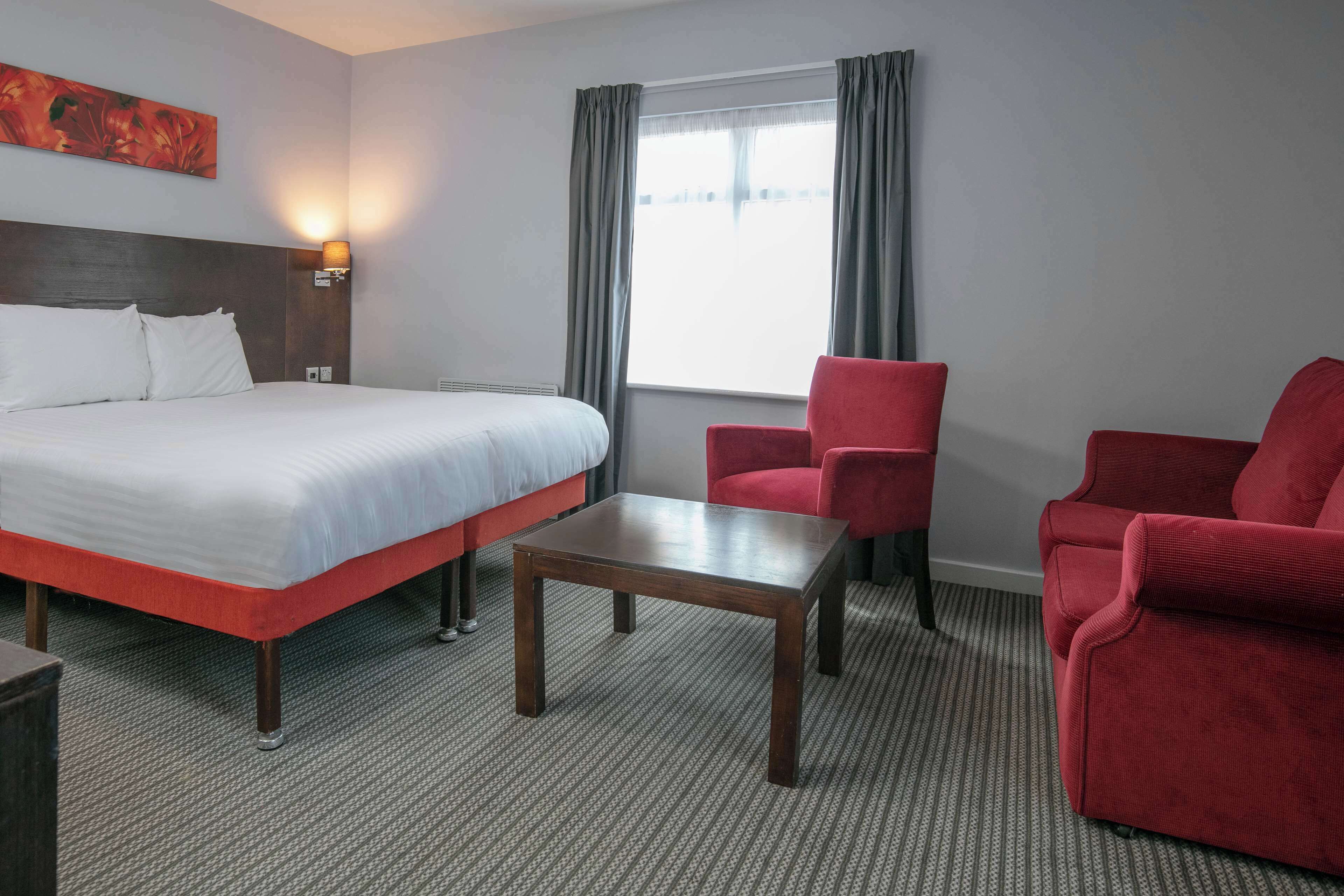 A hotel room with a bed, two red armchairs, a small table, a painting on the wall, and a window with curtains.